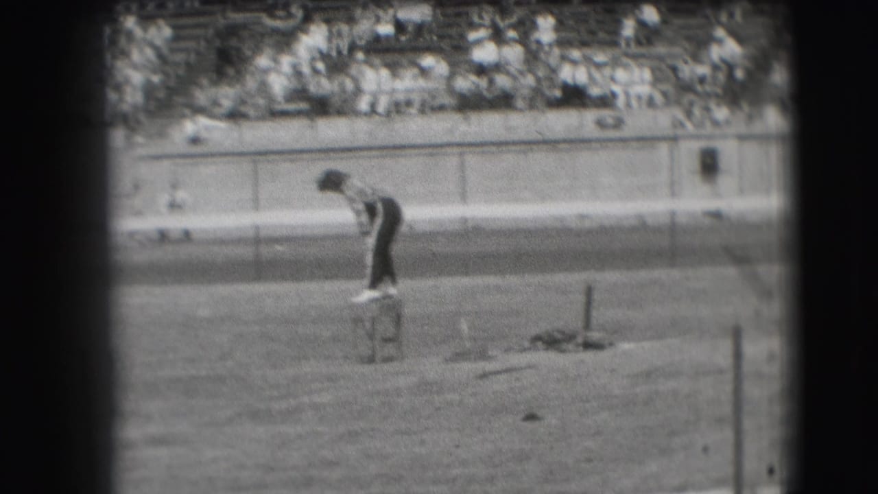 Man standing on a small stool near a racetrack with spectators in bleachers behind him.