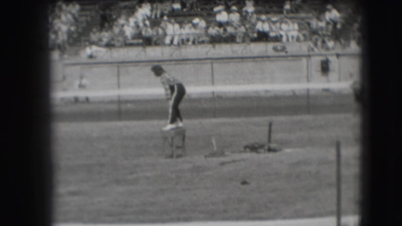 Man on a stool near the racetrack as blurred spectators sit in the stands.