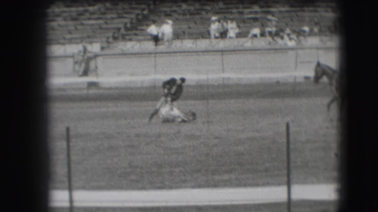 In the Denver rodeo arena, the cowboy leans over the roped animal on the ground with bleachers and spectators behind him.