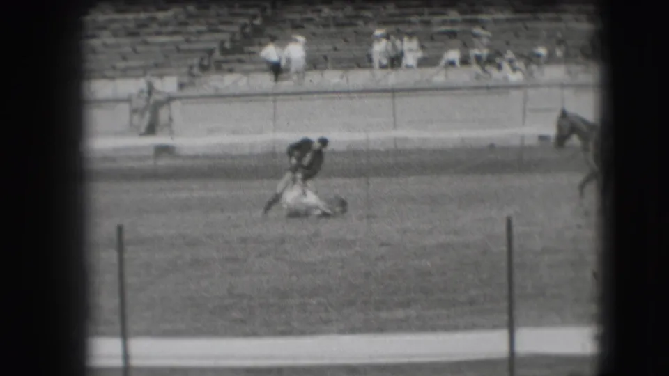 In the Denver rodeo arena, the cowboy leans over the roped animal on the ground with bleachers and spectators behind him.
