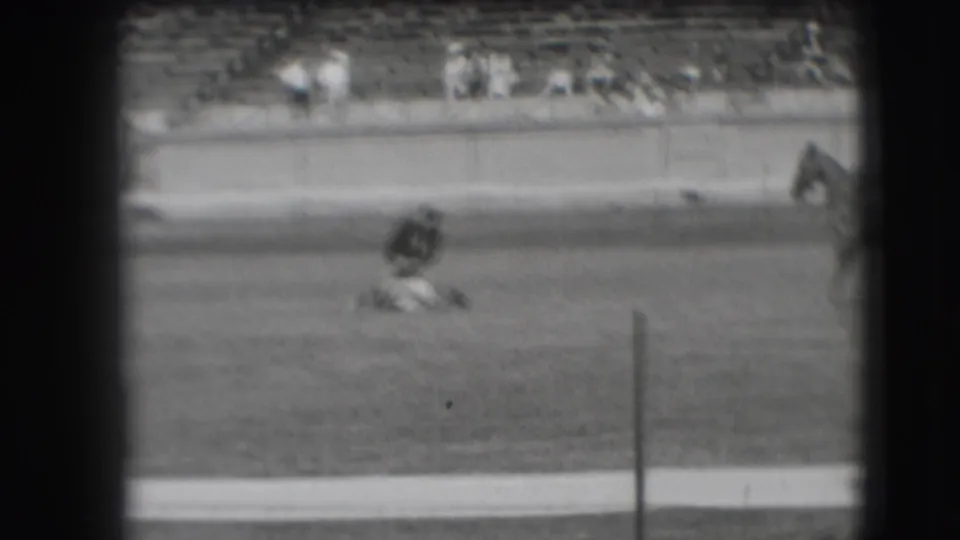 Soft 8mm frame shows the cowboy crouched low over the roped animal in the 1941 Denver arena, with spectators seated behind the rail.