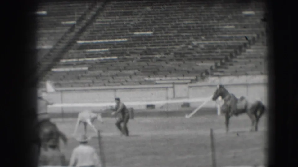 A ground handler bends toward a small animal as a mounted rider and another horse stand in the 1941 Denver rodeo arena.
