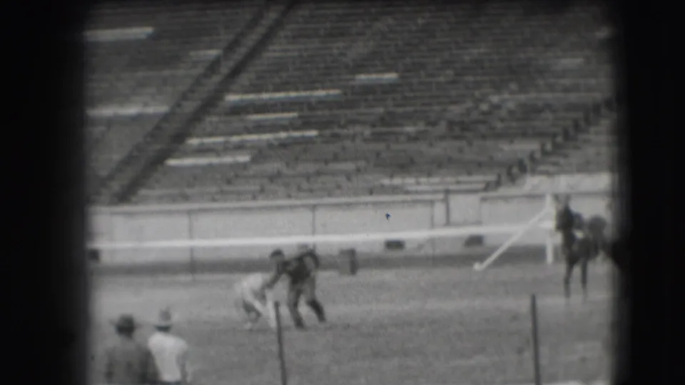 Two men appear to handle or steady a small animal in the Denver rodeo arena while a horse stands near the right edge of frame.