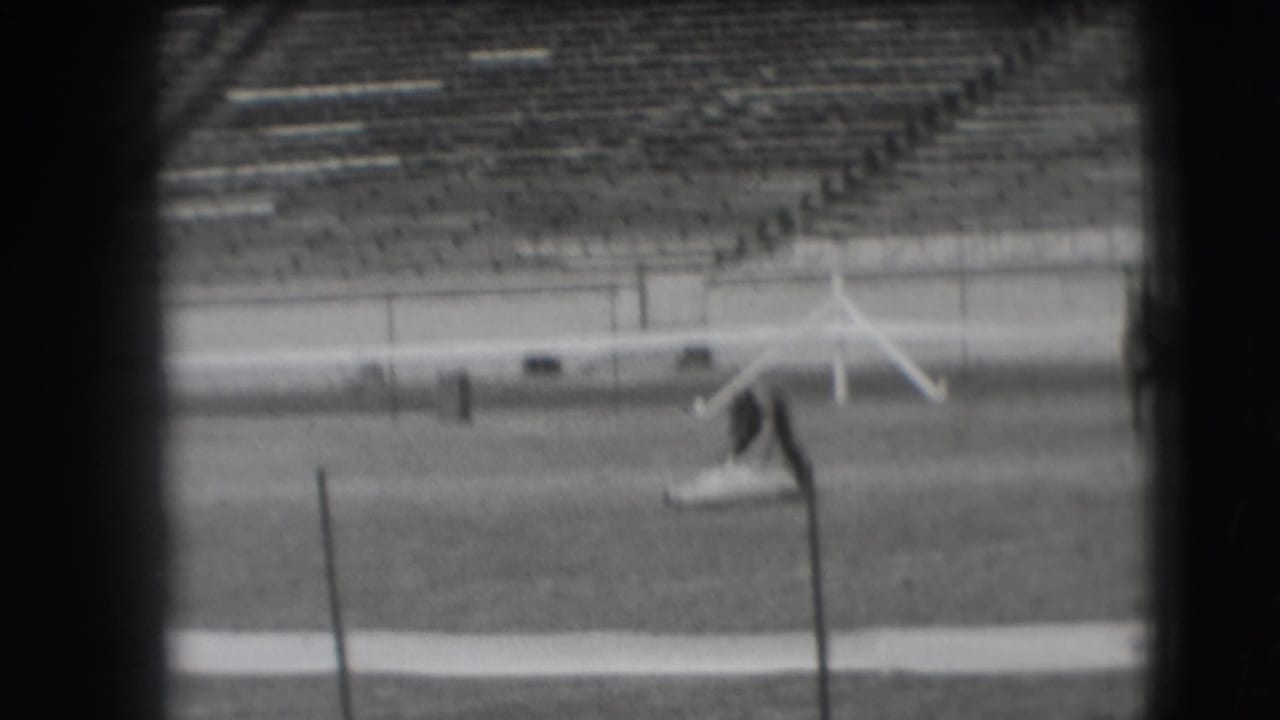 Mounted rider begins moving across a grass rodeo arena with empty stadium seating behind.