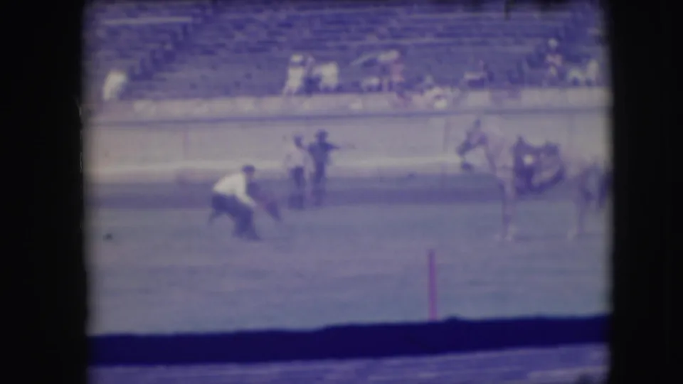 The contestant bends near the calf on the dirt arena floor while the horse stands to the side and spectators sit in the bleachers.