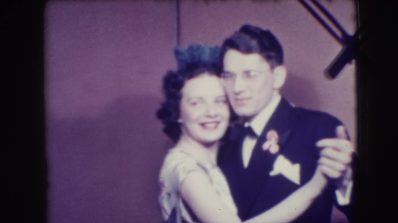 Well-dressed adult couple dancing close together against a plain wall.