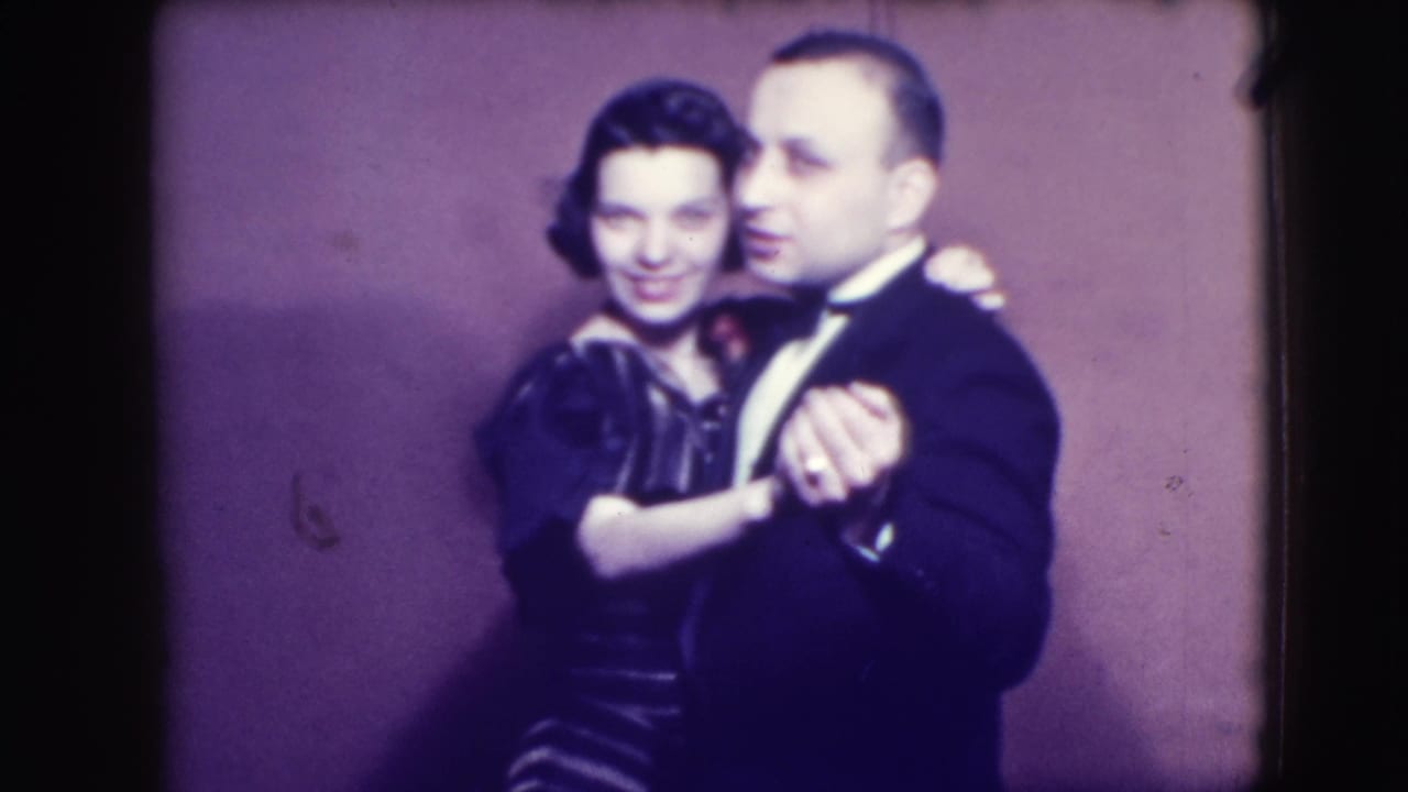 Adult man in tuxedo and adult woman in dark dress dancing closely before a plain purple backdrop.