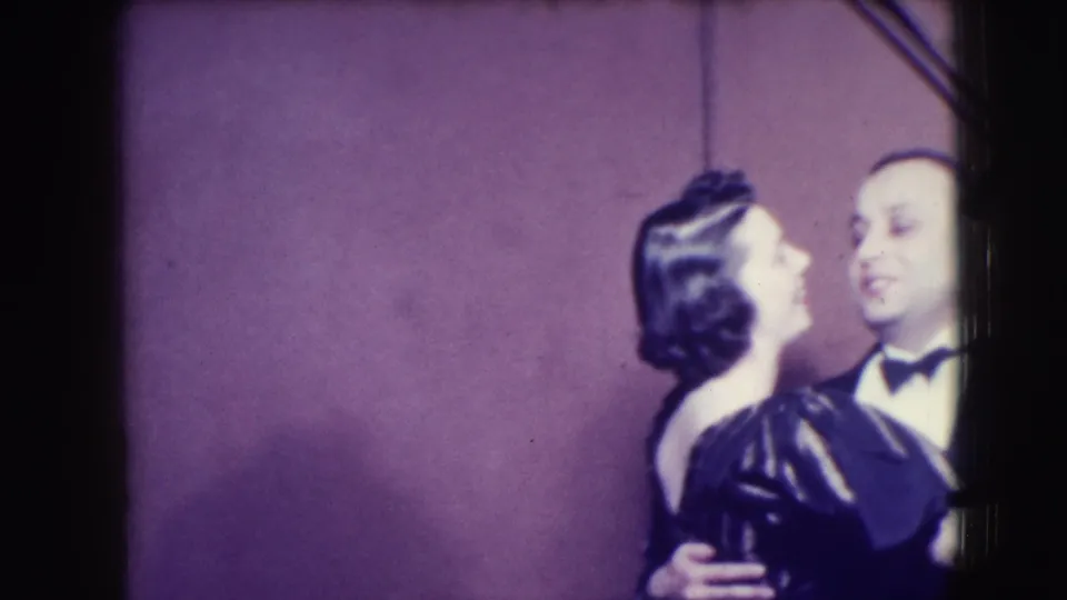 In 1941 Joliet home-movie footage, a woman in a dark dress and headpiece dances with a man in a tuxedo before a plain backdrop.