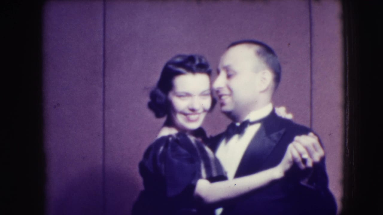 The couple smiles while dancing in formal clothes, with the woman facing the camera and the man turned slightly beside her.