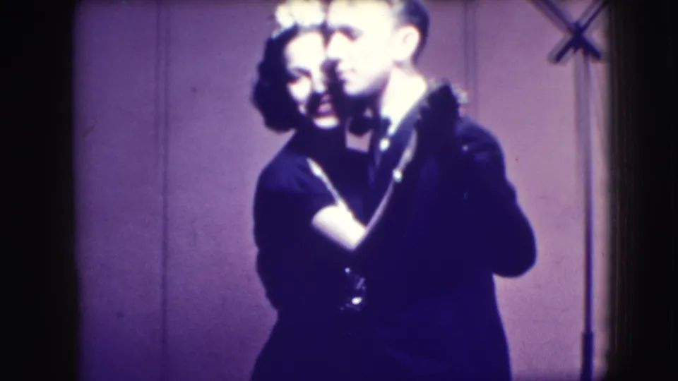 The couple remains in a close ballroom hold, partly shadowed, with a microphone stand or stage equipment visible near the backdrop.