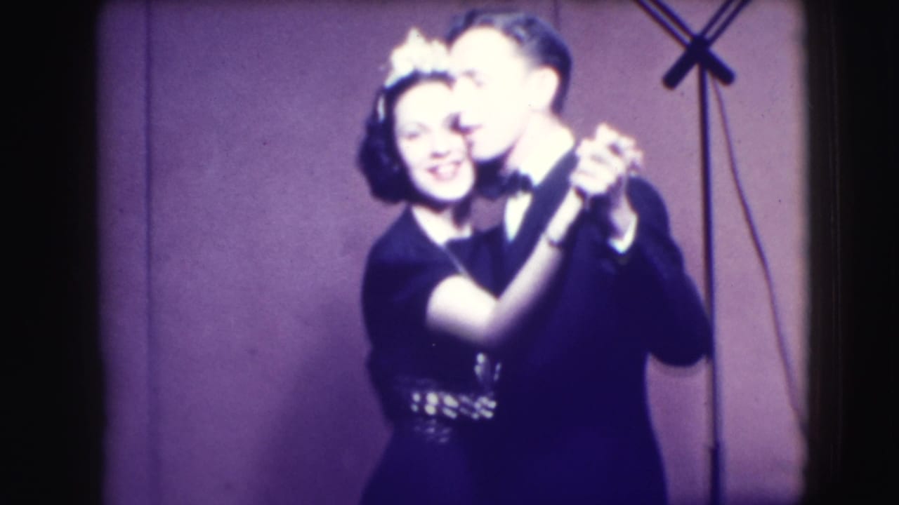 Smiling couple in formal attire dancing before a plain backdrop.