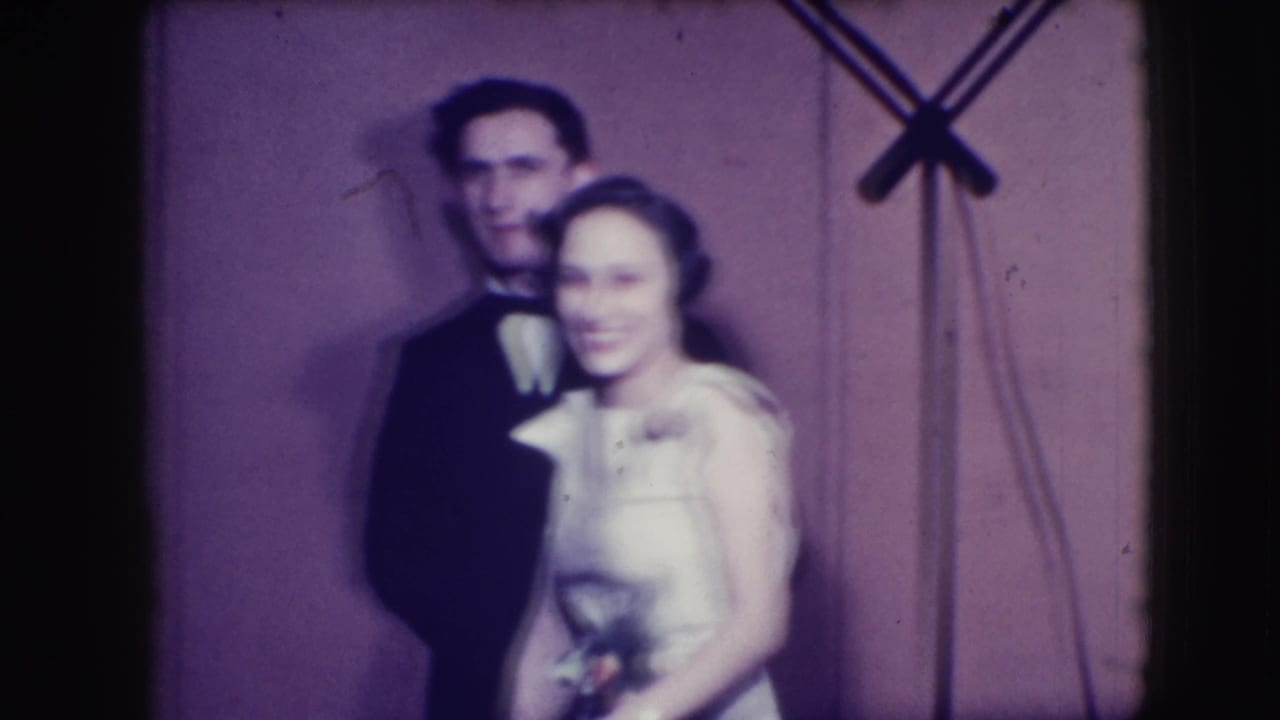 Young couple in formalwear standing close before a plain backdrop with a microphone stand nearby.