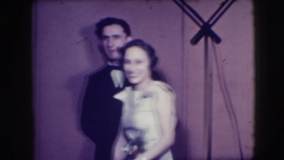 In 1941 Joliet home-movie footage, a young man in a dark tuxedo stands behind a woman in a light formal dress before a plain backdrop.