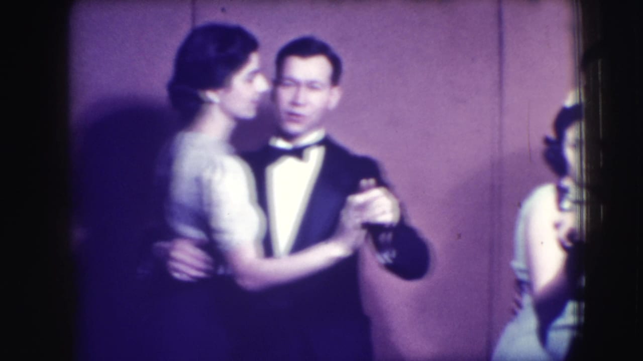 A woman in a light dress dances with a man in a tuxedo near a purple wall.