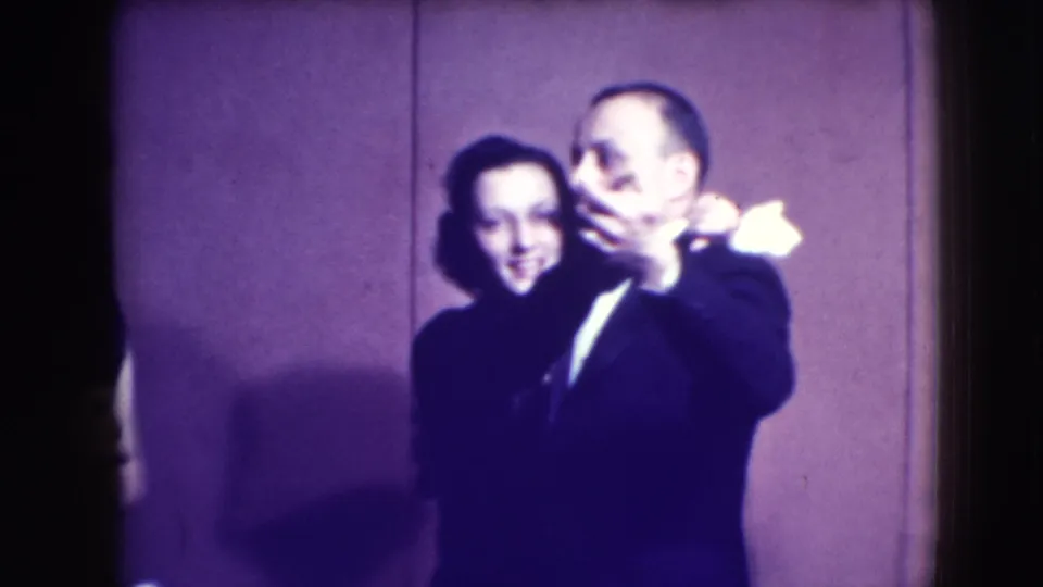 A formally dressed adult couple slow dances close to the camera, with another partial figure visible at the left edge.