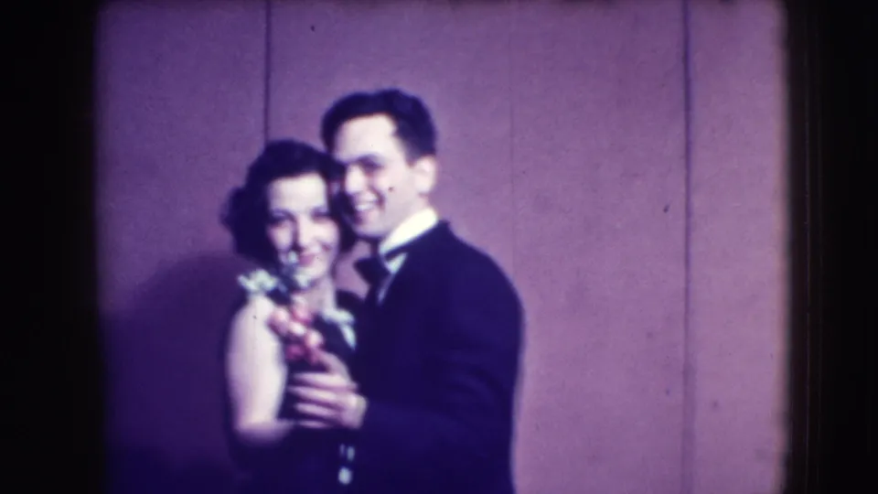 A smiling adult couple in formal clothing slow dances, with the woman's corsage visible in the 1941 Joliet home-movie footage.
