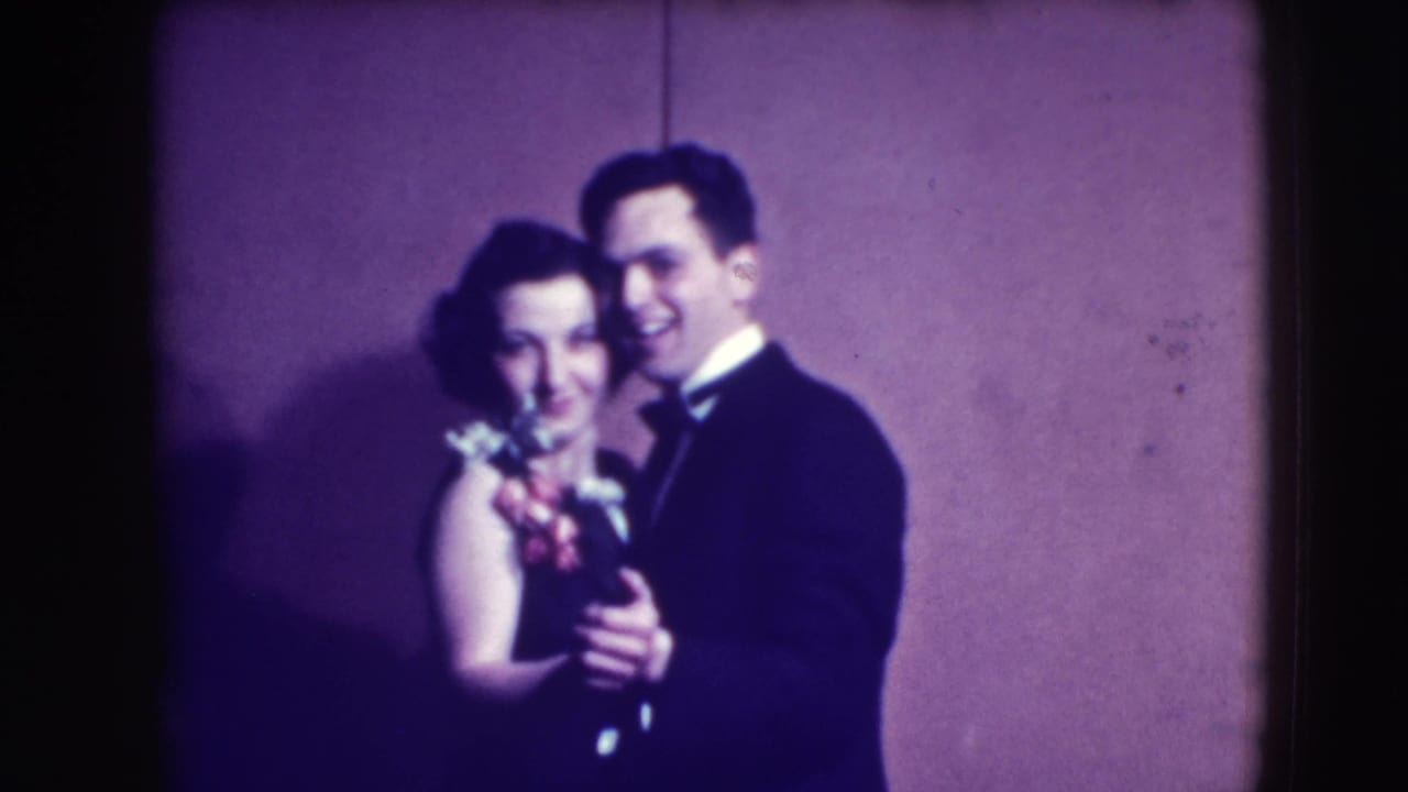 Well-dressed couple smiling and dancing against a plain wall.
