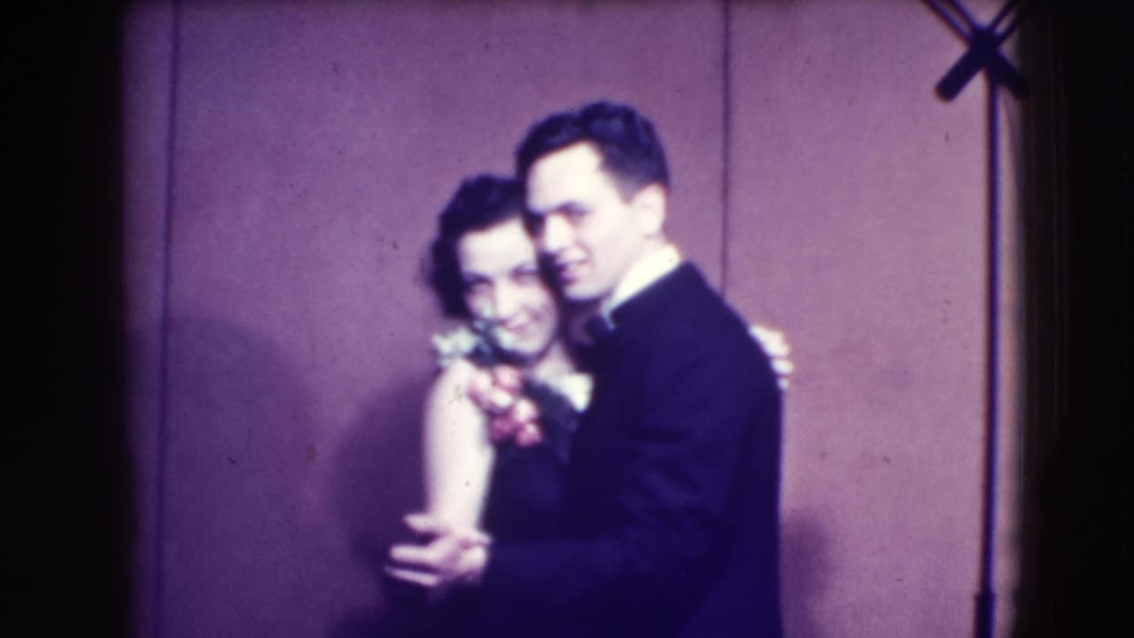 Couple in formal dress dances in front of a plain backdrop.