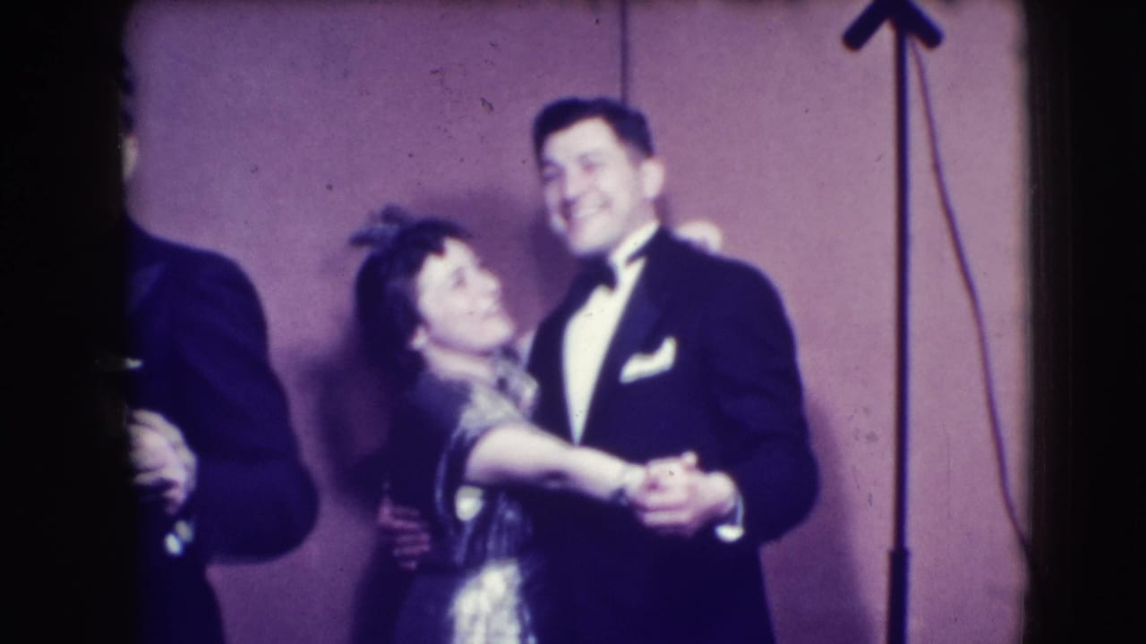 Man in tuxedo and woman in dress dance together beside microphone stands.