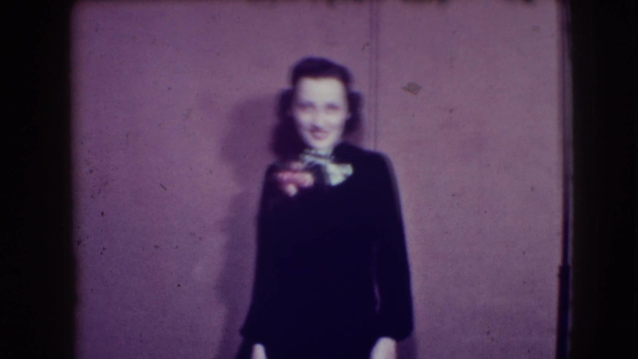 Woman in a dark dress stands smiling against a plain wall.
