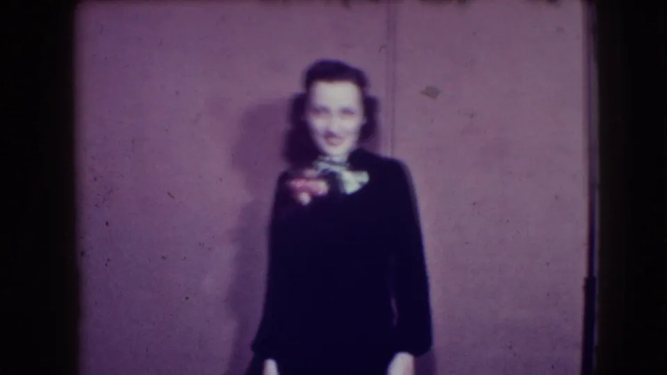 In this 1941 Joliet home-movie frame, a woman in a dark formal dress smiles toward the camera against a plain wall.