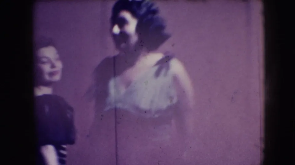 One woman in a pale gown stands in profile while another woman is partly visible behind her, both seen in soft-focus home-movie color.