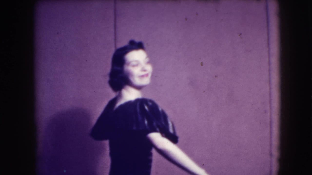 Brunette woman in a dark puffed-sleeve dress turns and smiles on a plain stage.