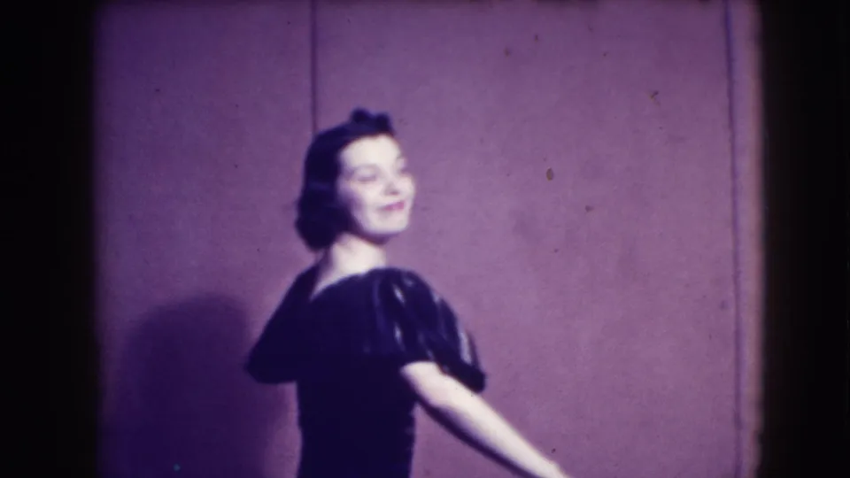 In 1941 Joliet home-movie footage, a brunette woman poses in a dark puffed-sleeve dress before a plain stage wall.