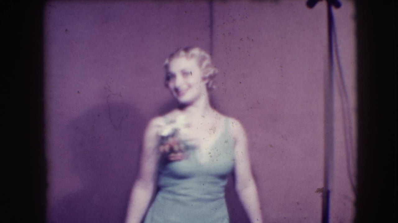 Blonde woman in a light sleeveless dress smiles on a stage while holding a small bouquet.