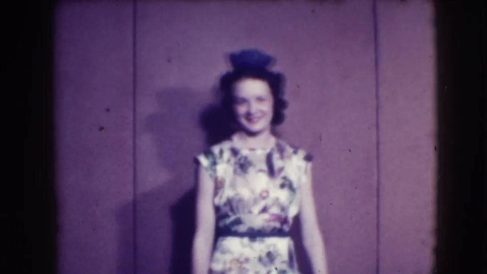 A woman in a floral dress, dark belt, and hat smiles on a Joliet stage in 1941 archival home-movie footage.