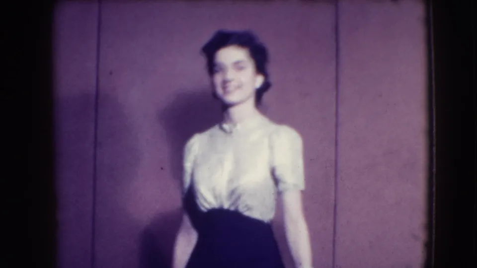 An adult woman in a white blouse and dark skirt faces the camera while modeling against the plain backdrop in the 1941 Joliet footage.
