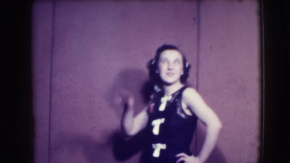 Smiling woman in 1941 Joliet home-movie footage poses in a dark sleeveless dress with white lettering and a hair flower.