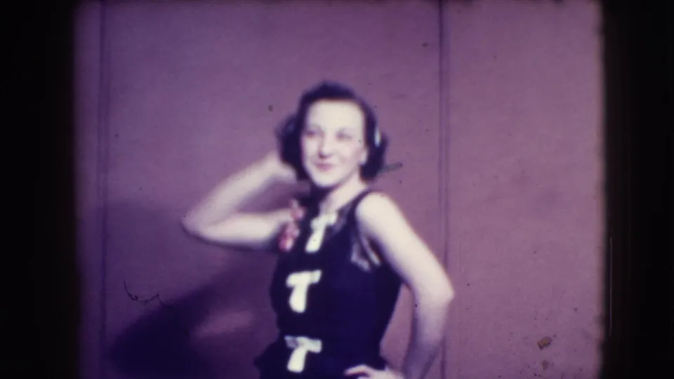 Young woman in 1941 Joliet footage smiles in a dark sleeveless dress, with one hand near her styled hair and flower accessory.