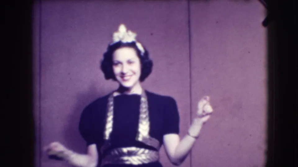 A woman in a dark dress with bright trim smiles and lifts both hands while posing against the purple-paneled wall.