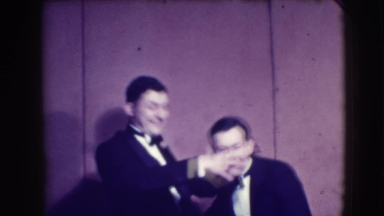 Two men in tuxedos laugh while one covers the other's eyes.
