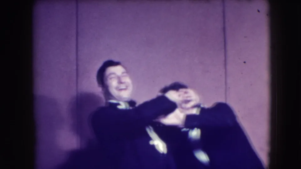 At the formal gathering, a tuxedoed man playfully covers a woman's eyes while both lean together and laugh.