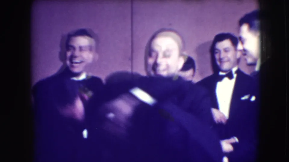 Several tuxedoed men gather in front of a plain wall, smiling and laughing in the Joliet formal-event home movie.