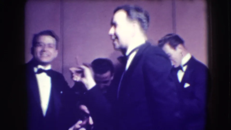 Men in formal tuxedos appear to joke and gesture in a Joliet, Illinois, 1941 home-movie frame, with faces visible and a plain wall behind them.