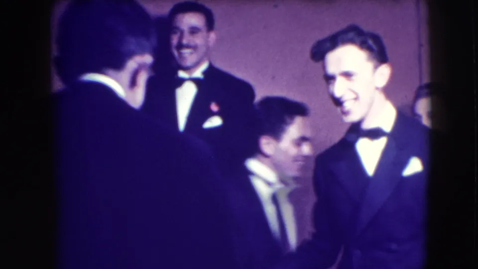 A 1941 Joliet home-movie frame shows tuxedoed men laughing and moving closely together during an indoor formal gathering.