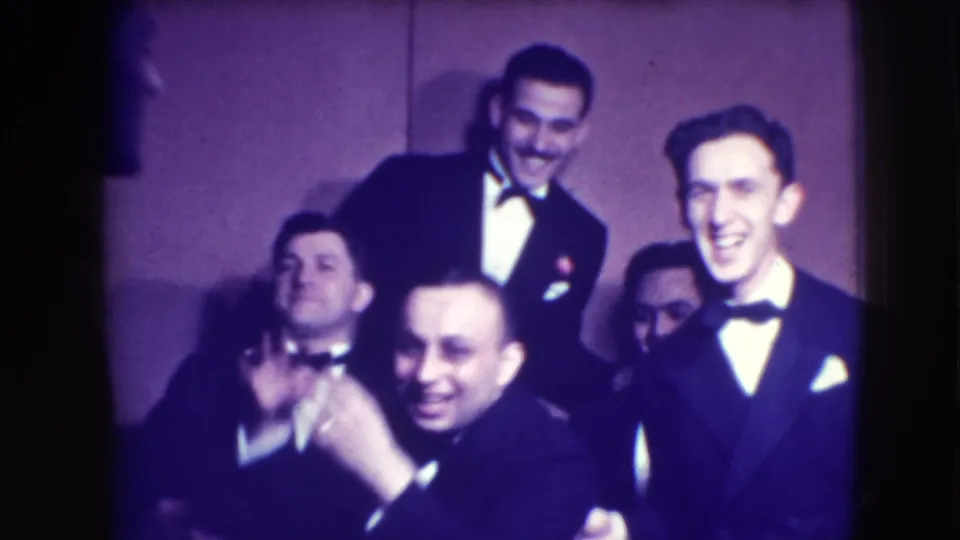 Tuxedoed men smile and pose toward the camera in a 1941 Joliet, Illinois, home-movie frame from an indoor formal gathering.