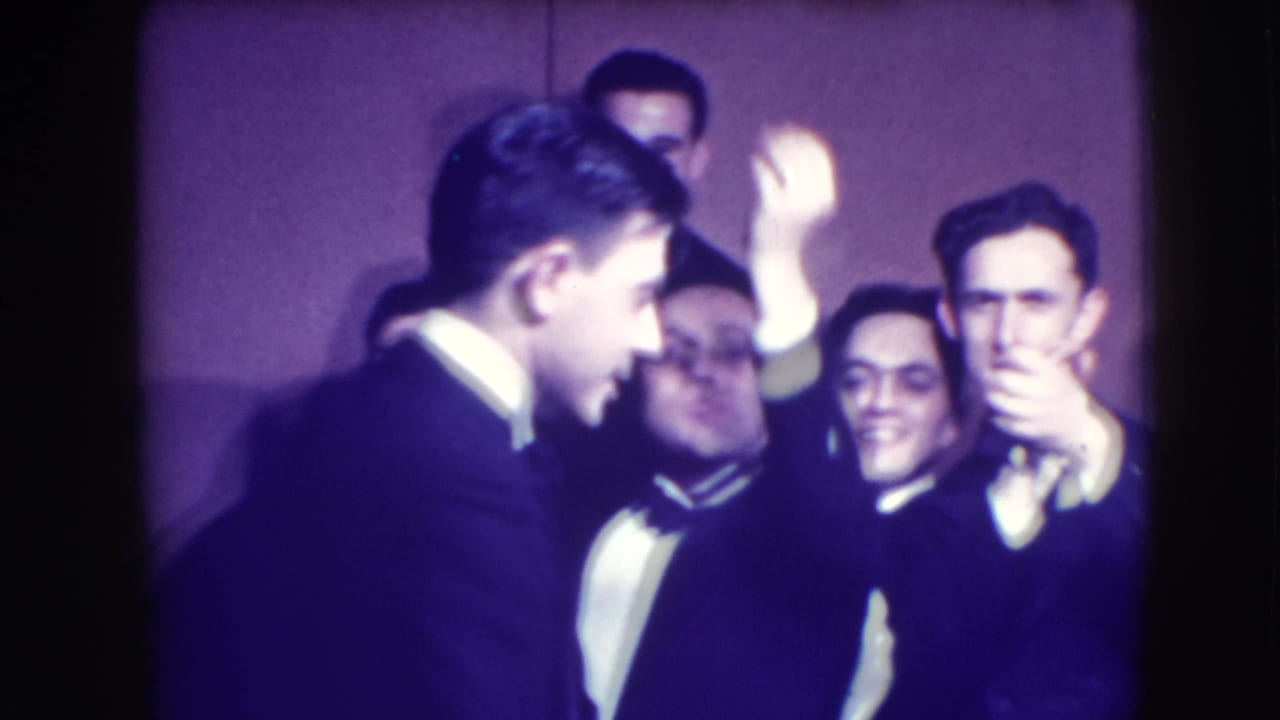 Men in tuxedos gather indoors, with one man in profile and others smiling behind him.