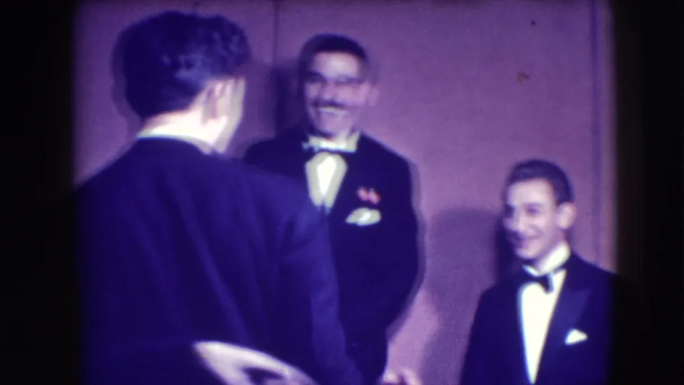 Formalwear-clad men stand against a plain interior wall, with one man turned away and two others smiling in the 1941 Joliet footage.