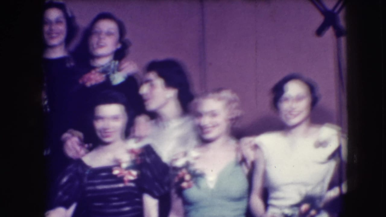 Group of young women in formal dresses and a man in a tuxedo pose indoors.
