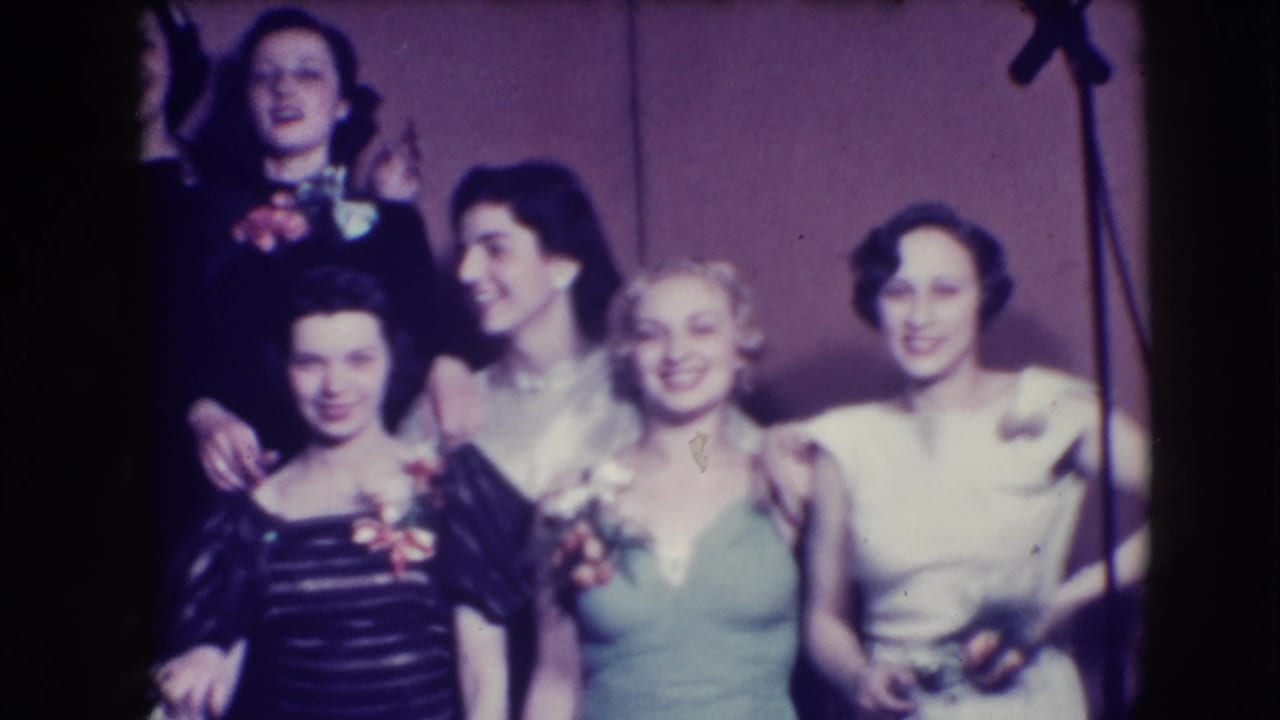 Women in formal gowns and a man in a tuxedo stand together, smiling toward the camera.