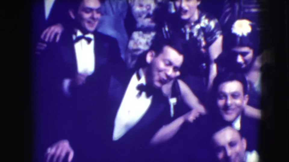 A man in a tuxedo leans toward the camera as other adults in formal dress stand behind him at the Joliet gathering.