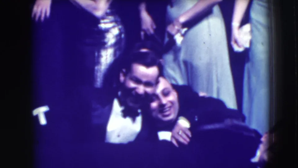 Two tuxedoed men lean together in a close foreground pose while other formally dressed guests stand behind them in the Joliet home movie.