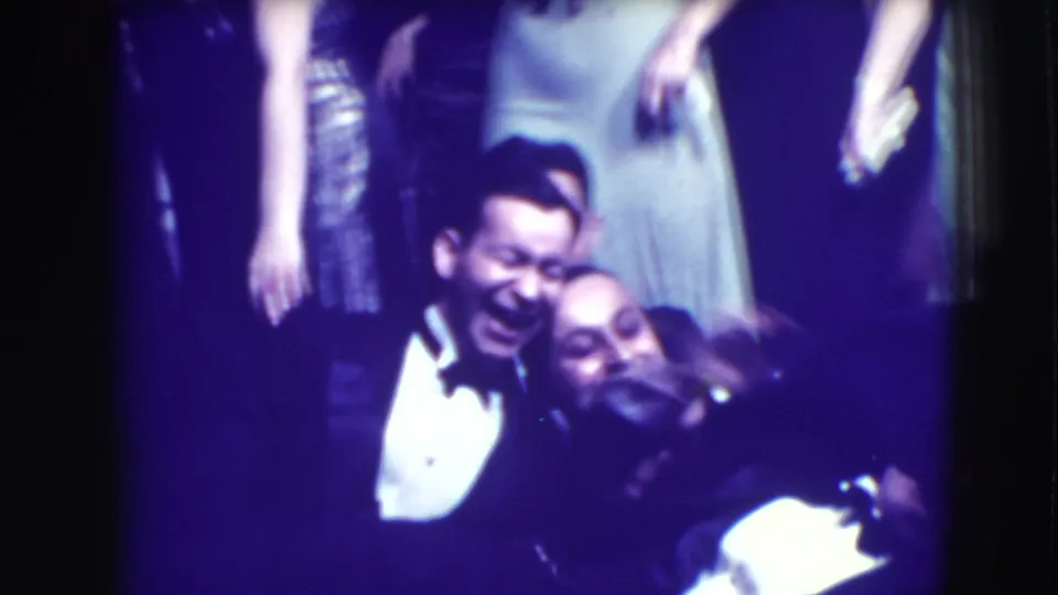 In 1941 Joliet home-movie footage, formally dressed adults laugh and lean close together under dim purple-hued light.