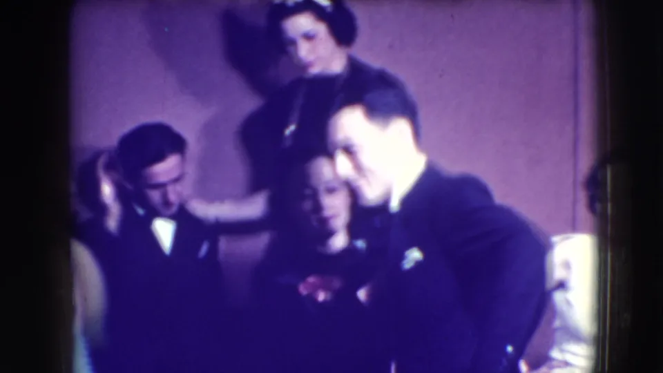 Adults in tuxedos and dresses gather closely at a Joliet formal party, with several faces visible in soft 8mm focus.