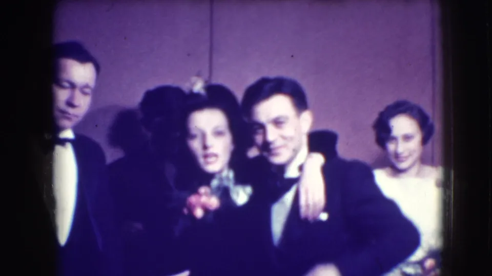 A woman wearing a corsage stands between tuxedoed men, with another woman behind them in 1941 Joliet party footage.
