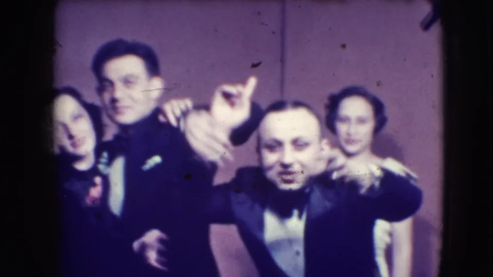 A woman and men in formal dress pose animatedly for the Joliet home camera, with hands raised and faces close to the lens.